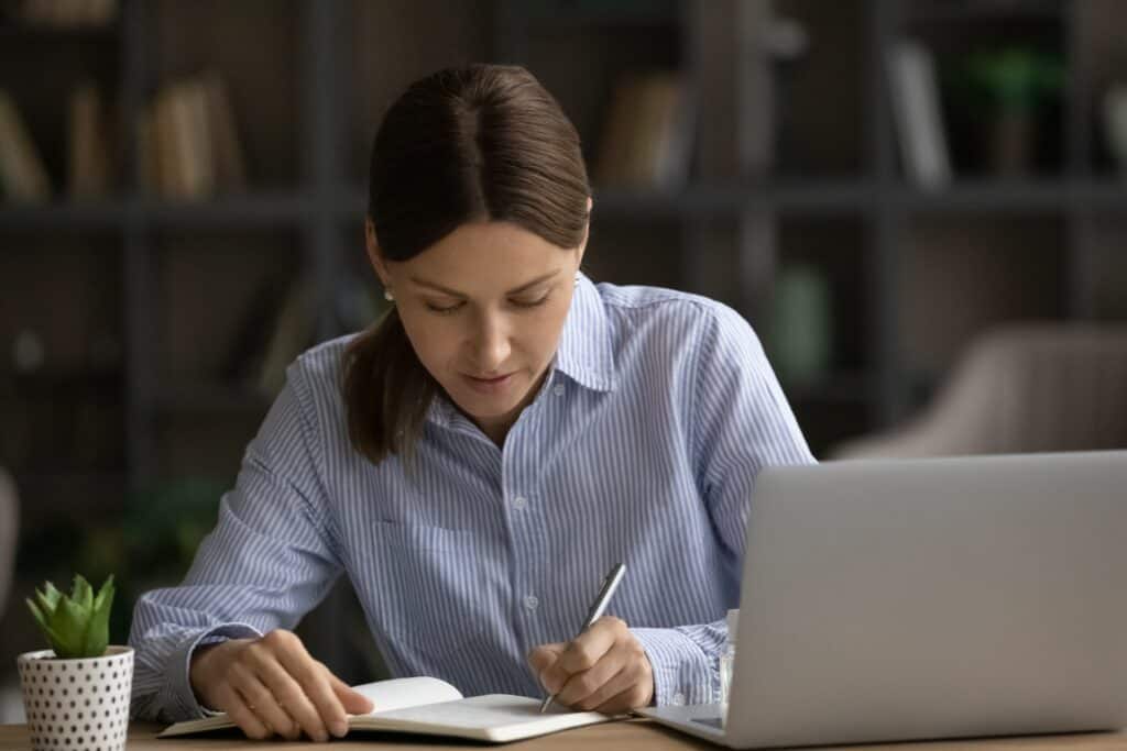 concentrated,serious,young,woman,sit,at,desk,near,laptop,jotting